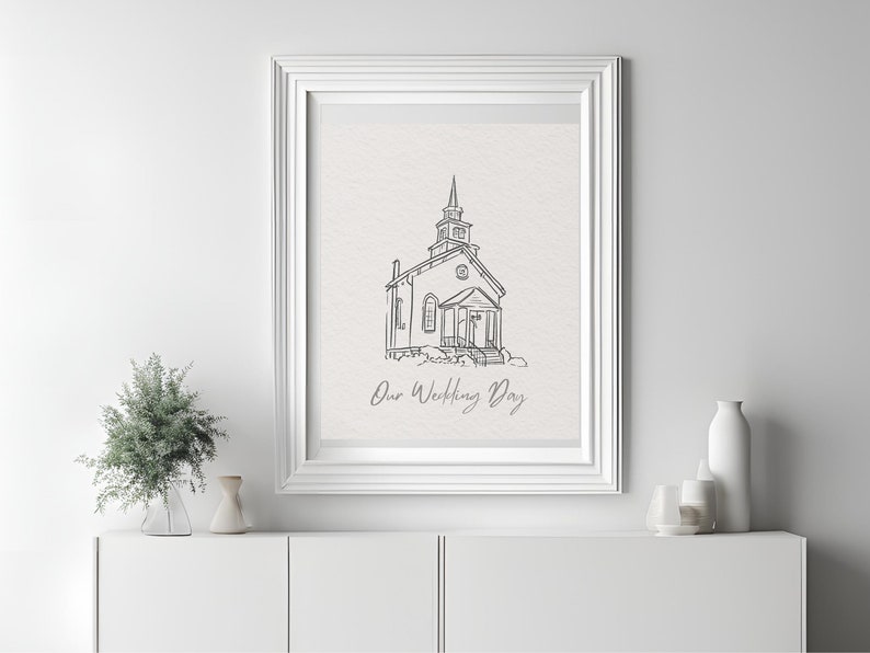 Op de afbeelding: Een zwart-witte lijntekening van een kerk met de tekst "Our Wedding Day" eronder. De tekening is ingelijst in een wit frame en hangt aan een witte muur.
