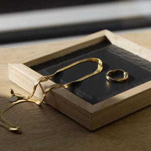 Könnte beinhalten: Eine goldfarbene Fischgrätenkette und ein kleiner, strukturierter goldfarbener Ring liegen in einem quadratischen Holztablett mit schwarzem Boden. Das Tablett hat einen hellen Holzrahmen und eine dunkle, leicht reflektierende Innenfläche.