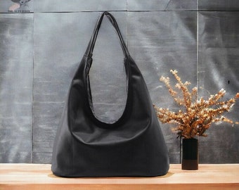 May include: A solid black hobo bag with a wide, integrated shoulder strap and a curved top opening. The bag features a smooth, matte finish and a spacious, unstructured body. A small logo with the text THE ALTY BAGS is visible in the top left corner.