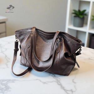 May include: A brown leather tote bag with a shoulder strap and top handles. The bag has a textured appearance and a zipper closure. The Alty Bags logo is visible in the upper left corner.