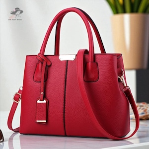May include: A red handbag with a textured finish, featuring two top handles and a detachable shoulder strap. The bag has a rectangular shape with a metal accent on the front and gold-coloured hardware. The brand name "THE ALTY BAGS" is visible in the top left corner.