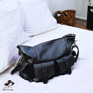 Puede incluir: Un bolso de hombro negro con acabado liso, un compartimento principal con cremallera y dos bolsillos frontales con detalles de correas verticales. El bolso incluye herrajes de metal plateado, enganches laterales y el texto THE ALTY BAGS.