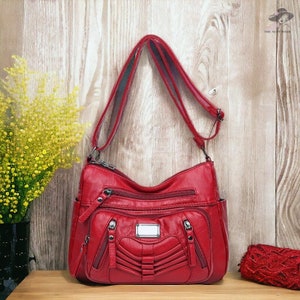Puede incluir: Un bolso bandolera de color rojo brillante con una correa larga ajustable, múltiples compartimentos exteriores con cremallera y una placa metálica rectangular central. El bolso tiene una superficie texturizada con detalles de costuras horizontales.