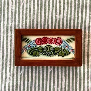 May include: A rectangular ceramic tile framed in dark brown wood, featuring a central design of red circular shapes, green lobed leaves, and blue floral accents on a cream background. The tile displays the word GOOD in stylized red lettering.