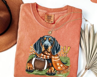 May include: A peach-colored t-shirt with a black and brown dog wearing an orange and white scarf and holding a football. The dog is surrounded by a floral design and the text "YUM" is in the upper right corner of the image.