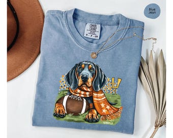 May include: A blue jean t-shirt with a cartoon illustration of a black and tan dog wearing an orange and white scarf and holding a brown football. The dog is sitting in front of a yellow and white football goal post. The text "COMFORT COLORS" and "S - P - CH" are printed on the shirt.