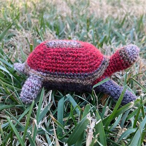 May include: A crocheted turtle toy with a red, brown, and purple shell. The turtle is lying on its back in a bed of green grass.