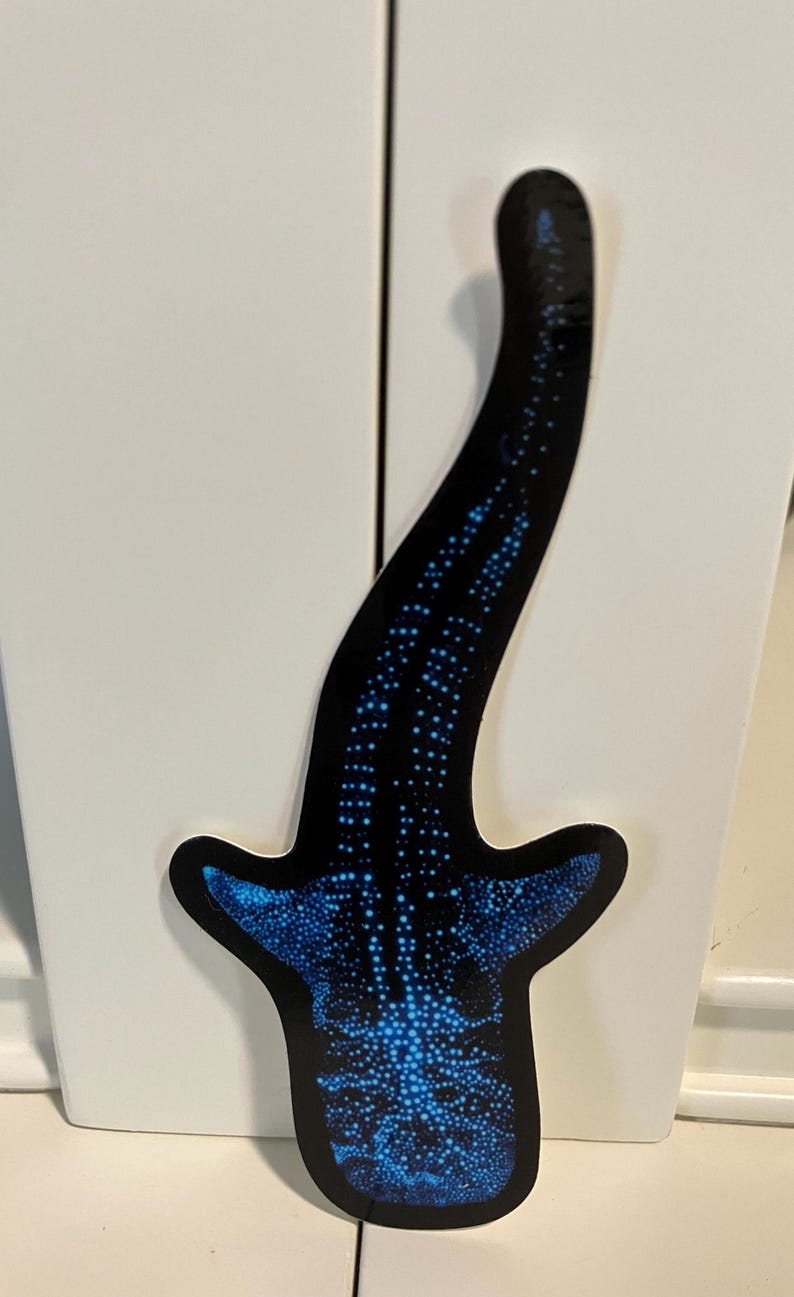 May include: A blue and black whale shark sticker with a white outline. The sticker is on a white surface.