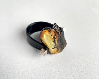 May include: A black ring with a silver accent and a large amber stone. The amber stone is a mix of brown, yellow, and white.