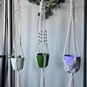 May include: Three off-white macrame plant hangers suspended from a metal rod. Each hanger features intricate knotted patterns, a wooden ring at the top, and a tassel at the bottom. They hold a white ribbed pot, a green pot, and a purple pot.