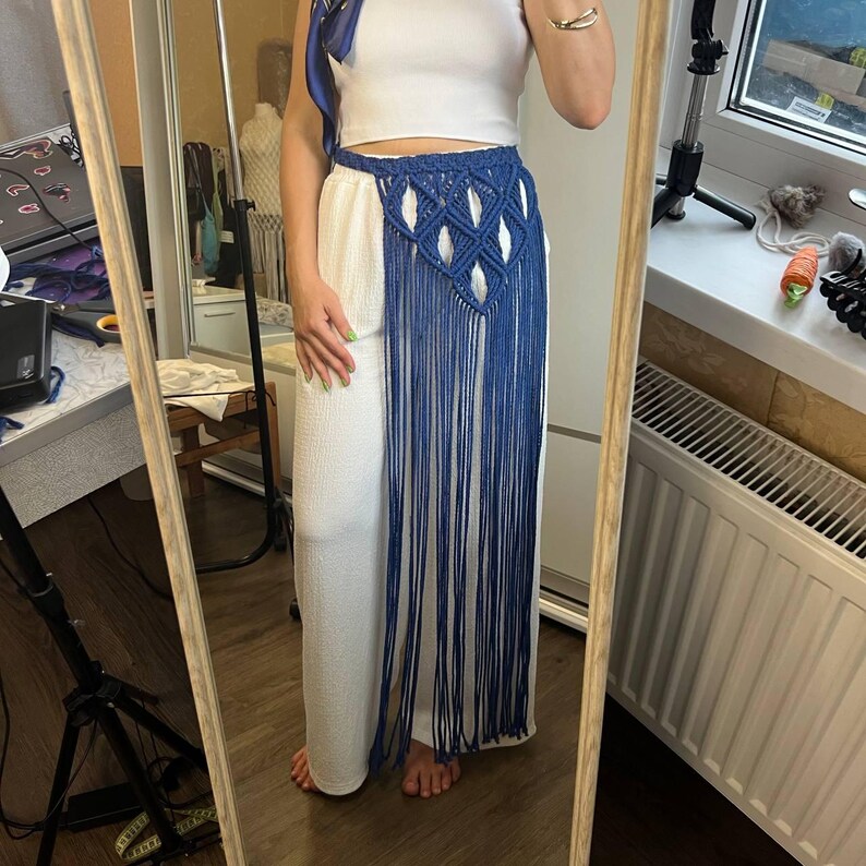 May include: A blue macrame waist accessory featuring a diamond-patterned knotted belt section and long, flowing fringe that extends downward. The piece is worn over white trousers and a white top, showcasing the intricate cord work and texture.