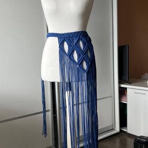 May include: A deep blue macrame skirt featuring a woven waistband with a diamond-shaped knotted pattern and long, flowing fringe that extends downward. The garment is displayed on a white mannequin, showcasing the intricate cord work and length.