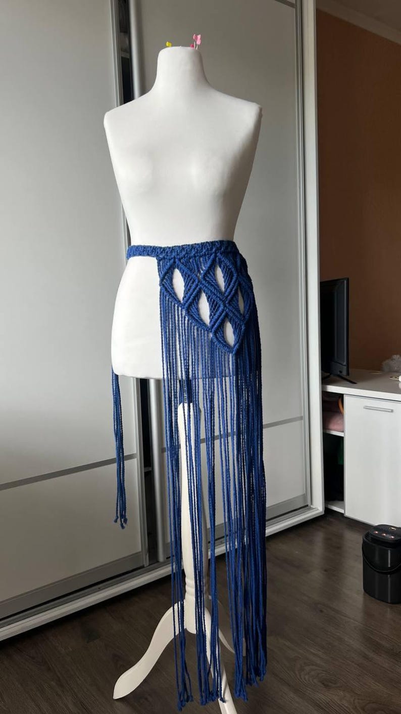 May include: A deep blue macrame skirt featuring a woven waistband with a diamond-shaped knotted pattern and long, flowing fringe that extends downward. The garment is displayed on a white mannequin, showcasing the intricate cord work and length.