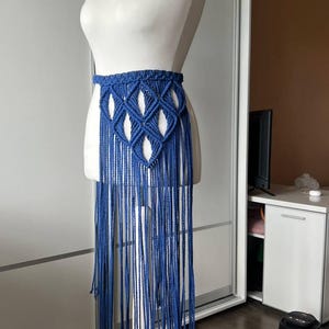 May include: A blue macrame belt featuring a woven diamond pattern at the waist and long, dangling fringe that extends downward. The belt is displayed on a white mannequin torso, showcasing the intricate knotted cord construction and texture.