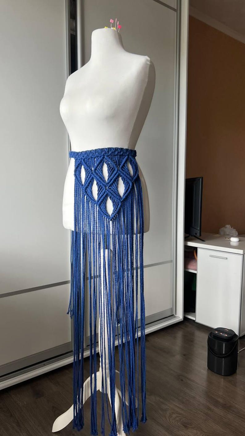 May include: A blue macrame belt featuring a woven diamond pattern at the waist and long, dangling fringe that extends downward. The belt is displayed on a white mannequin torso, showcasing the intricate knotted cord construction and texture.