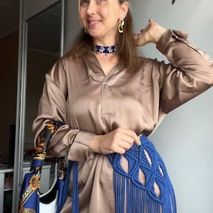 May include: A long-sleeved, button-down satin shirt in a light brown hue, paired with a blue macram&eacute; waist accessory featuring long, dangling fringe. The ensemble includes a blue and gold patterned silk scarf and a blue beaded choker necklace.