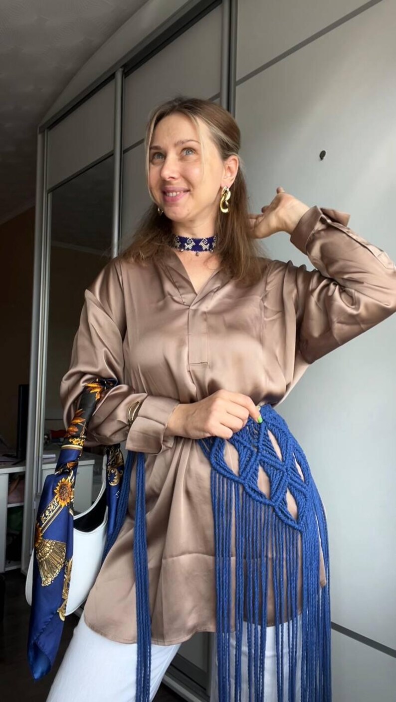 May include: A long-sleeved, button-down satin shirt in a light brown hue, paired with a blue macram&eacute; waist accessory featuring long, dangling fringe. The ensemble includes a blue and gold patterned silk scarf and a blue beaded choker necklace.