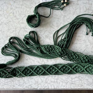 Peut inclure: Une ceinture en macramé vert foncé présentant un motif complexe de nœuds en forme de losange sur toute la bande principale. La ceinture comprend de longues franges en corde et une petite boucle circulaire ornée de deux perles en bois clair.
