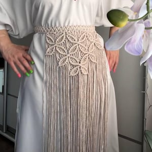 May include: A beige macrame belt featuring an intricate knotted floral pattern across the front section, with long, vertical fringe strands hanging down from the waist. The belt is worn over a plain white dress with a red string at the neckline.