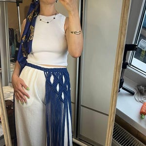 May include: A white sleeveless ribbed top paired with white wide-leg trousers featuring a dark blue macrame waist accessory. The accessory includes a knotted diamond pattern and long, flowing fringe that extends down the side of the trousers.