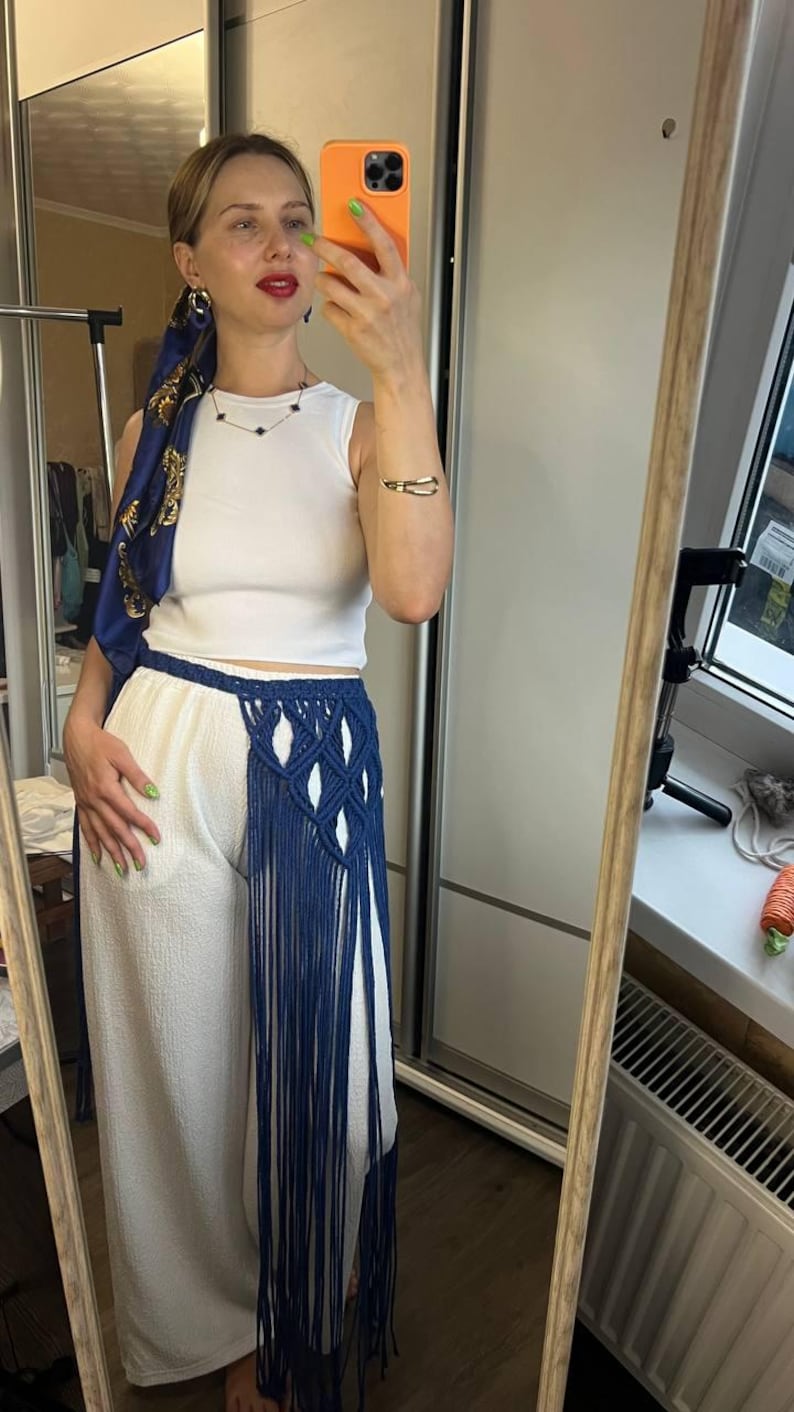 May include: A white sleeveless ribbed top paired with white wide-leg trousers featuring a dark blue macrame waist accessory. The accessory includes a knotted diamond pattern and long, flowing fringe that extends down the side of the trousers.