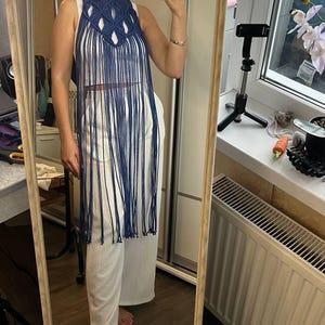 May include: A dark blue macrame halter top featuring an intricate knotted design at the neckline and long, flowing fringe that extends down to the waist. The garment is worn over a plain white sleeveless top paired with white wide-leg trousers.