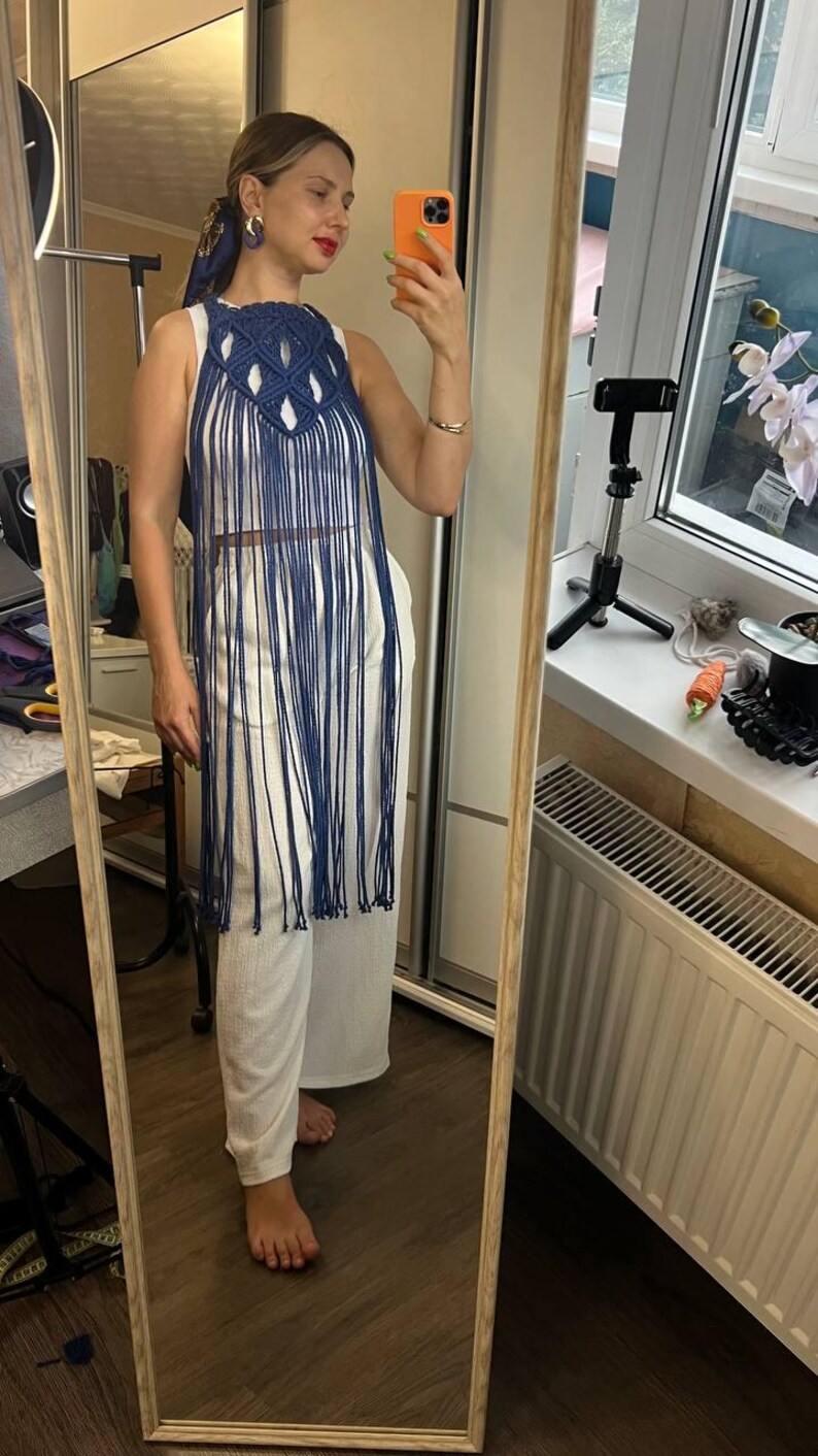 May include: A dark blue macrame halter top featuring an intricate knotted design at the neckline and long, flowing fringe that extends down to the waist. The garment is worn over a plain white sleeveless top paired with white wide-leg trousers.