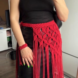 May include: A bright red macrame waist accessory featuring an intricate knotted diamond pattern and long, flowing fringe that extends downward. The piece includes a matching red macrame wristband, both crafted from thick cord with a textured finish.