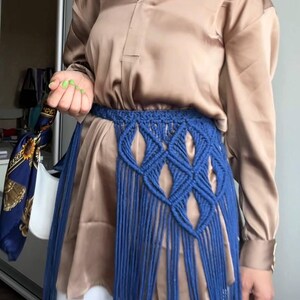 May include: A beige satin button-down shirt paired with white trousers and a blue macrame belt featuring a diamond-patterned weave and long fringe. A blue choker with white patterns and small silver bells is worn around the neck.