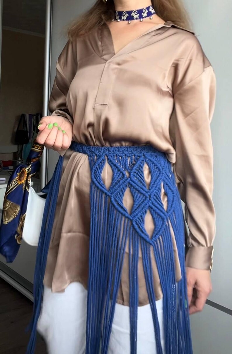 May include: A beige satin button-down shirt paired with white trousers and a blue macrame belt featuring a diamond-patterned weave and long fringe. A blue choker with white patterns and small silver bells is worn around the neck.