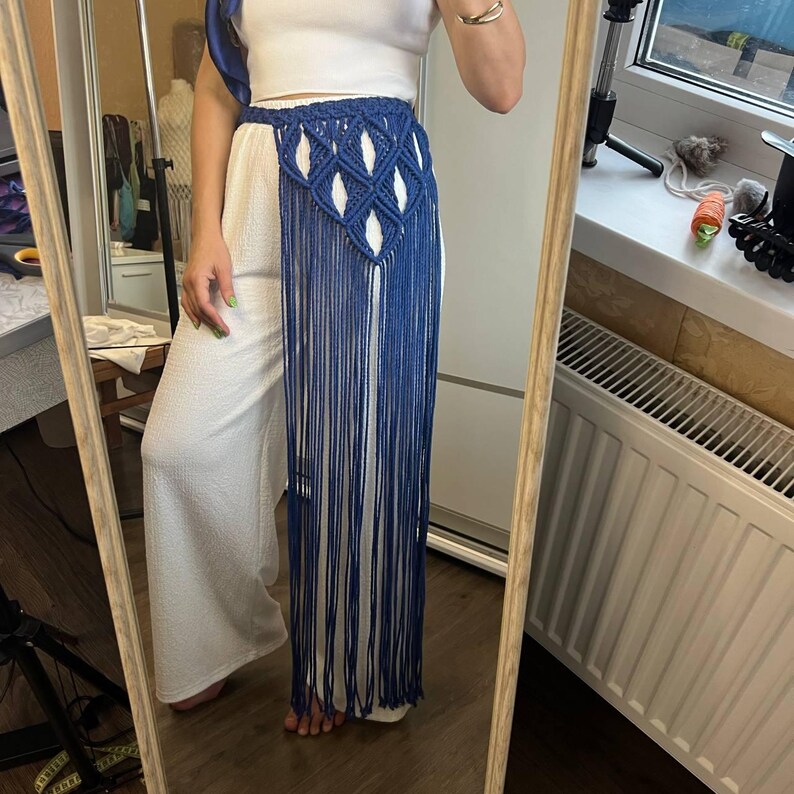 May include: A blue macrame waist accessory featuring a diamond-shaped knotted pattern at the top and long, flowing fringe that extends downward. The piece is worn over a white sleeveless top and white wide-leg trousers, creating a layered look.