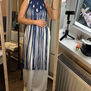 May include: A dark blue macrame overlay featuring a knotted geometric pattern at the chest and long, flowing fringe that extends to the knees. The piece is worn over a simple white sleeveless top and matching white wide-leg trousers.