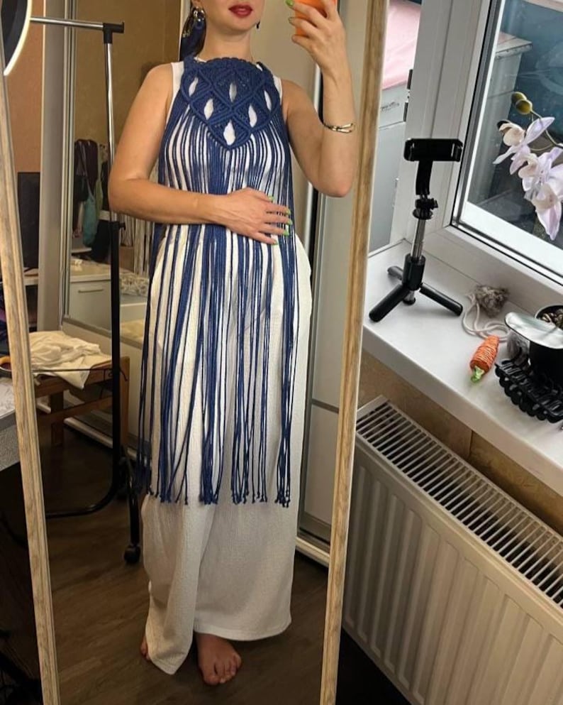 May include: A dark blue macrame overlay featuring a knotted geometric pattern at the chest and long, flowing fringe that extends to the knees. The piece is worn over a simple white sleeveless top and matching white wide-leg trousers.