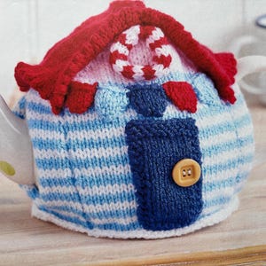May include: A white ceramic teapot covered by a knitted cosy shaped like a house. The cosy features a red roof, a blue and white striped body, a dark blue door with a yellow button, and a red and white striped circular accent on the roof.