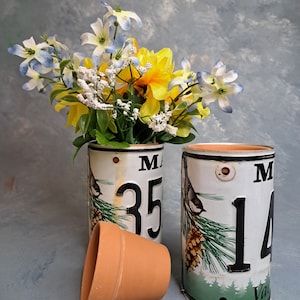 May include: Two white ceramic flower pots with a green and brown pine tree design and a small bird. The pots are decorated with the letters "M" and the numbers "35" and "14". The pots are filled with a bouquet of yellow and blue flowers.