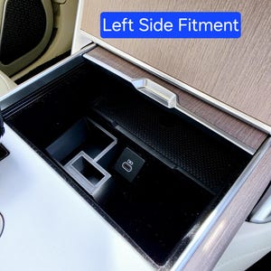 Könnte beinhalten: Autoinnenraumkonsole mit einem schwarzen Staufach und Holzoptik-Verkleidung. Die Konsole verfügt über einen USB-C-Anschluss und den Text "Left Side Fitment" in einem blauen Rechteck. Das Fach ist offen und zeigt sein Inneres.