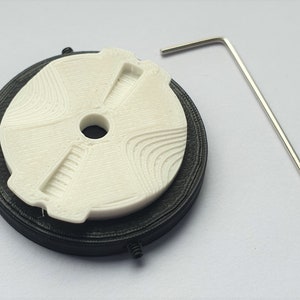 Peut inclure: Une pièce blanche imprimée en 3D avec une base noire et une petite clé Allen argentée. La pièce est circulaire avec un trou au centre et une série de crêtes sur la surface.