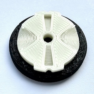 Puede incluir: Un objeto de plástico blanco impreso en 3D con un diseño en forma de cruz y un agujero en el centro. El objeto está montado sobre una base de plástico negro.