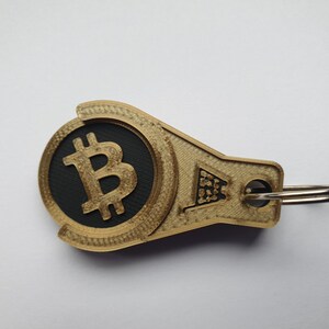 May include: A gold-colored keychain with a black circle featuring a gold Bitcoin symbol. The keychain has a silver ring.