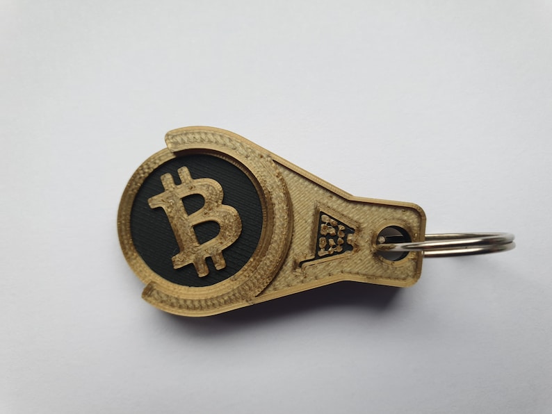 May include: A gold-colored keychain with a black circle featuring a gold Bitcoin symbol. The keychain has a silver ring.