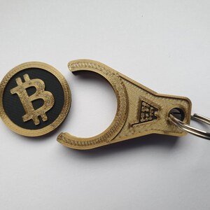 May include: A gold-colored keychain with a black and gold Bitcoin coin attached. The keychain has a loop for attaching to a bag or belt.