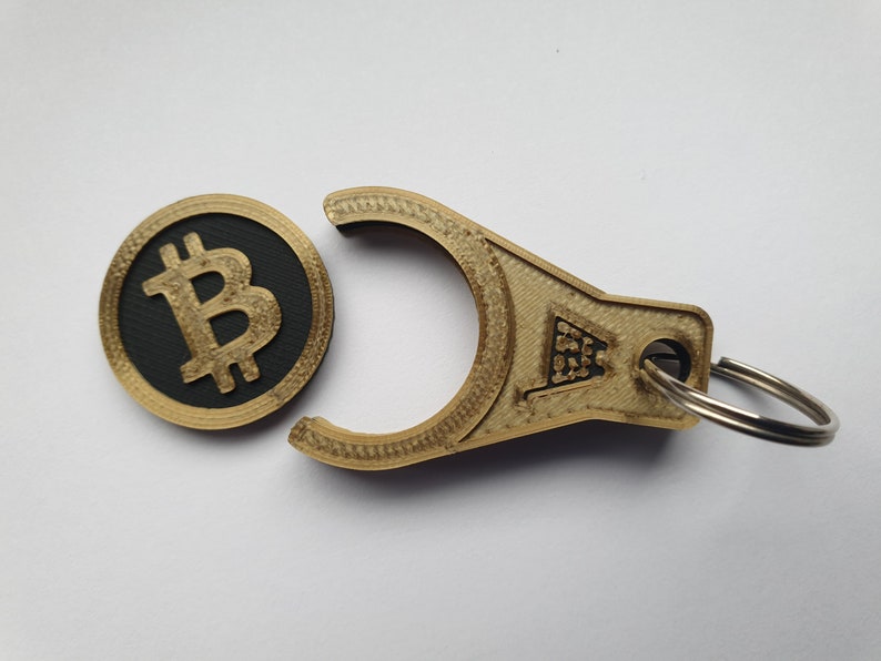 May include: A gold-colored keychain with a black and gold Bitcoin coin attached. The keychain has a loop for attaching to a bag or belt.
