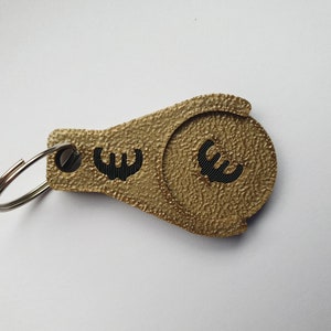 May include: Gold-colored keychain with a coin holder featuring a black euro symbol. The keychain has a silver ring.
