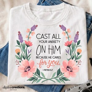 Op de afbeelding: Een wit t-shirt met een bloemenkransontwerp rondom de zwarte en koraalkleurige tekst: CAST ALL YOUR ANXIETY ON HIM BECAUSE HE CARES for you, 1 PETER 5:7. De krans bestaat uit roze, oranje en paarse bloemen met groene bladeren.