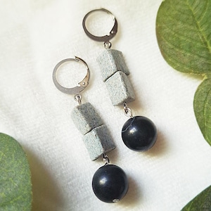 May include: A pair of dangle earrings with silver leverback closures. Each earring features three grey, cube-shaped beads and a black, spherical bead. The earrings are displayed on a white surface with green leaves.