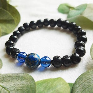 May include: A beaded bracelet featuring black onyx beads and blue jasper and glass beads. The bracelet is strung on an elastic cord. The bracelet is surrounded by green leaves.