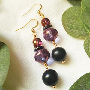 May include: Dangle earrings with gold-coloured hooks. Each earring has dark purple, brown, and black beads. The beads are connected by gold-coloured wire, creating a stylish accessory for any occasion.