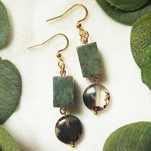 May include: A pair of dangle earrings with gold-colored hooks. Each earring features a rectangular green stone, a clear and brown circular bead, and a small gold-colored bead at the bottom. The earrings are set against a white background.