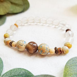 以下が含まれることがあります： さまざまな色と素材のビーズで作られたブレスレット。ブレスレットは、茶色、ベージュ、青の色合いの透明、つや消し、模様のビーズが特徴です。ブレスレットは、緑の葉を背景に白い表面に表示されています。