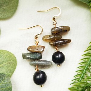 May include: Gold-coloured drop earrings with stacked, oblong stones in shades of grey, green, and brown, with a black bead at the bottom. The earrings have a simple hook design.